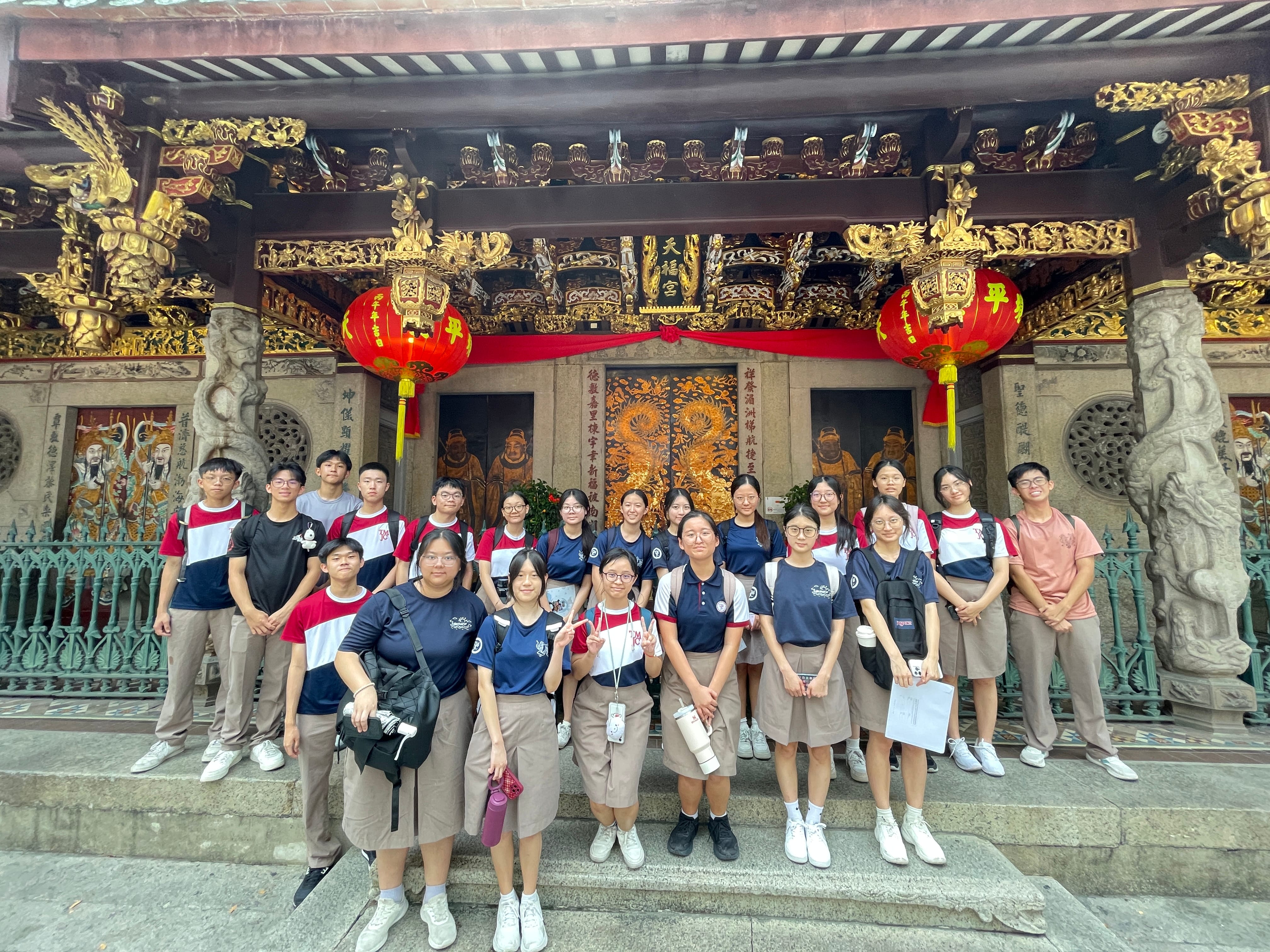 A group of about 20 students in school uniforms pose for a photo in front of an ornately decorated Chinese temple with red lanterns and gold carvings.
